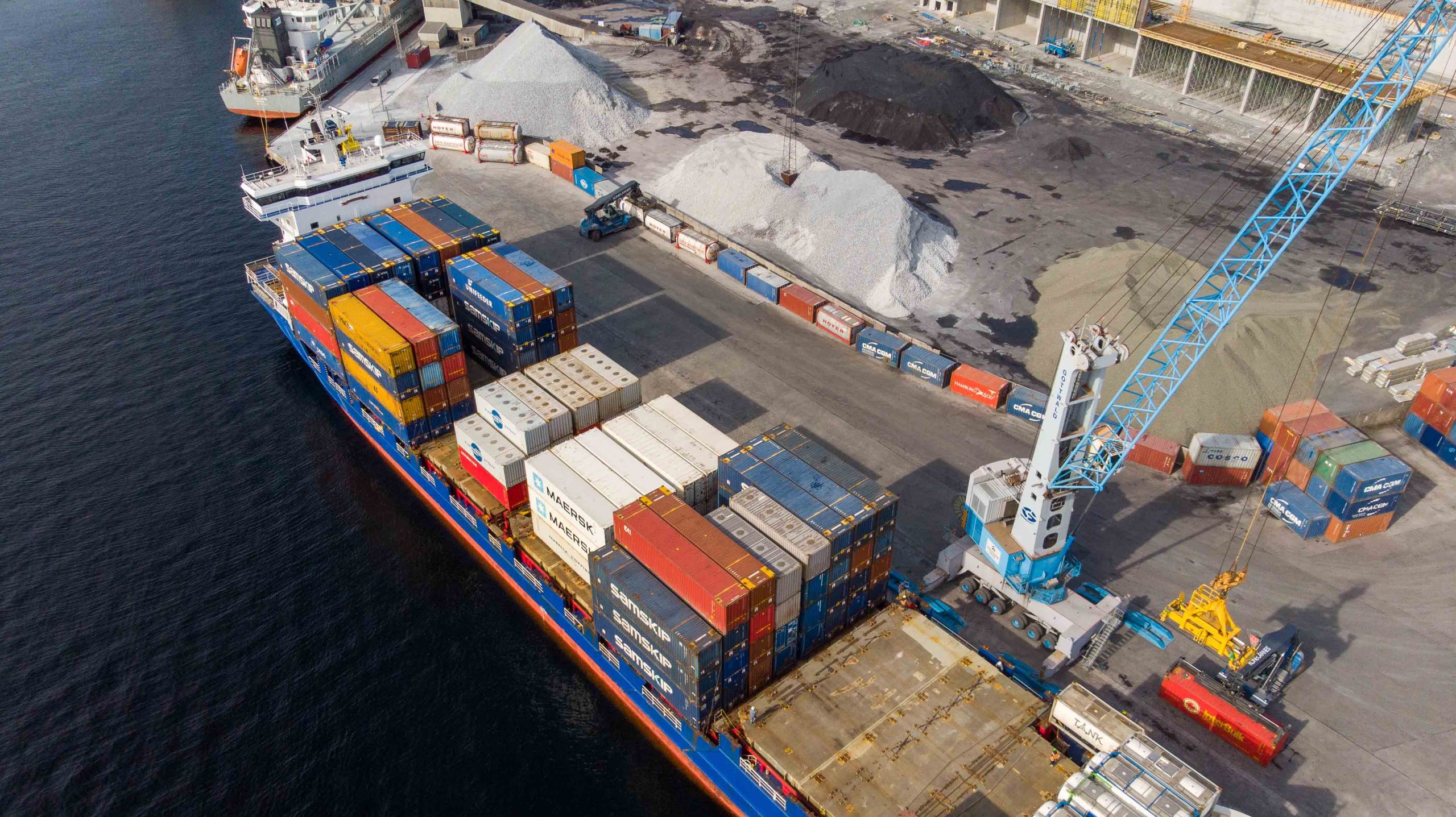 Skip fylt med containere ligger til kai. En mobil havnekran losser skipet for containere.